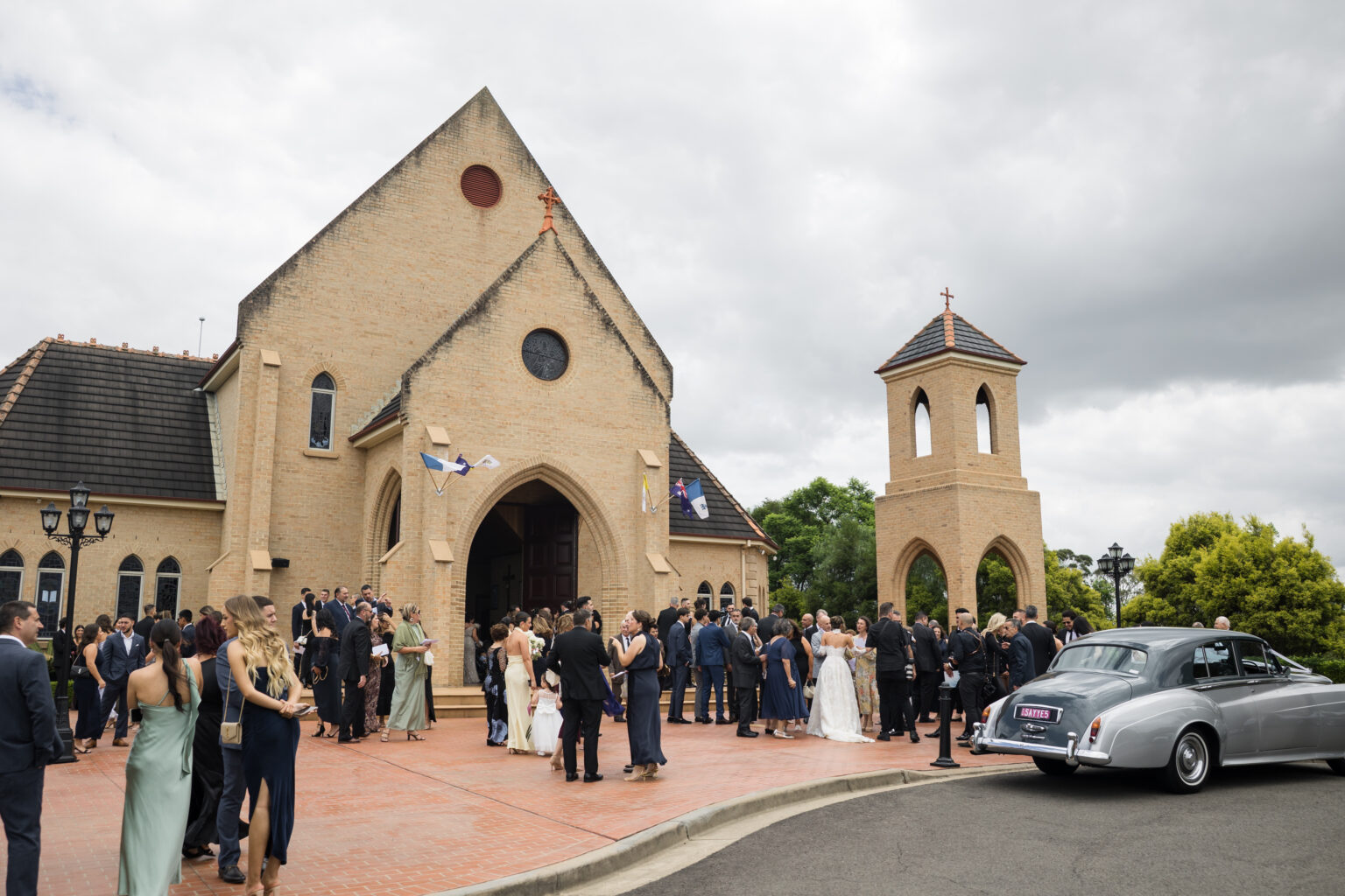 Wedding guests in formal attire gather outside a brick church with a bell tower on a cloudy day.