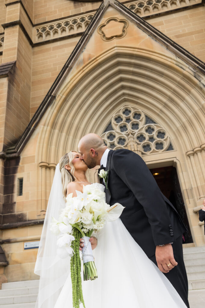 Bride and groom kiss outside a stone church with a large arched doorway and ornate window details, the bride holding a white bouquet.