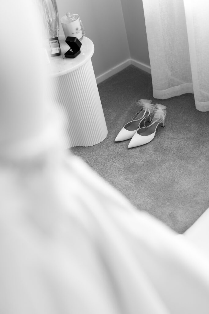 White pointed-toe bridal heels with bow details on a carpet near a sheer curtain, beside a round table with a jewelry box and perfume.