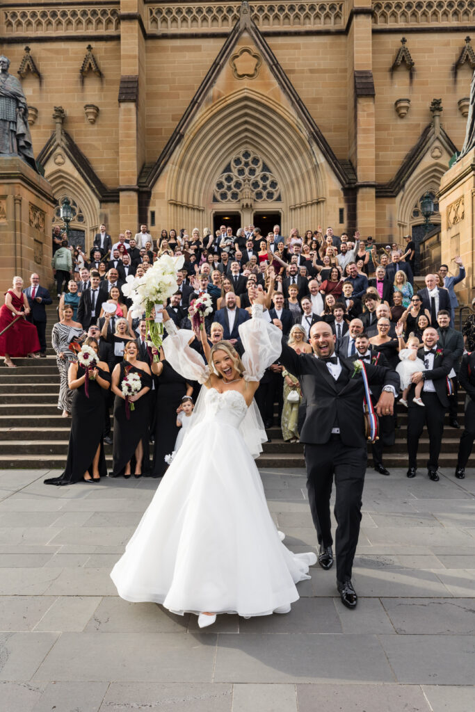 Bride in a white wedding dress leads a large group of guests down stone steps of a gothic church, everyone cheering and posing for photos.
