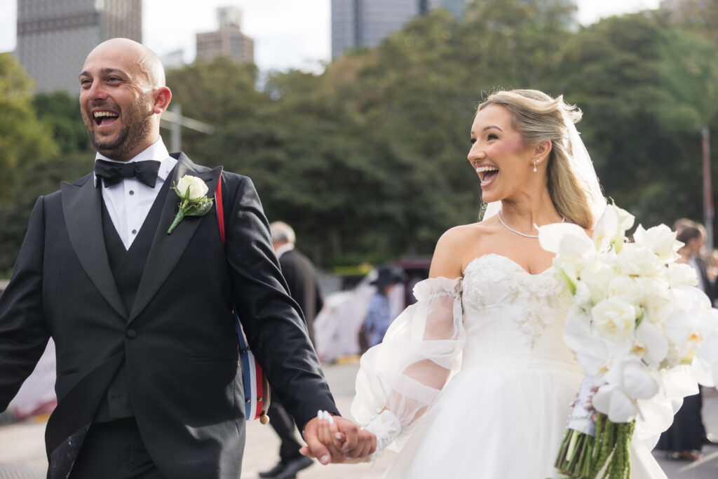 Bride and groom laughing while holding hands outdoors at their wedding, bride in a white gown with a large bouquet nearby.