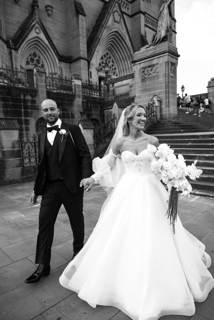 Black-and-white wedding photo of a groom in a tuxedo and a bride in a white gown walking hand in hand outside a gothic stone church and steps.