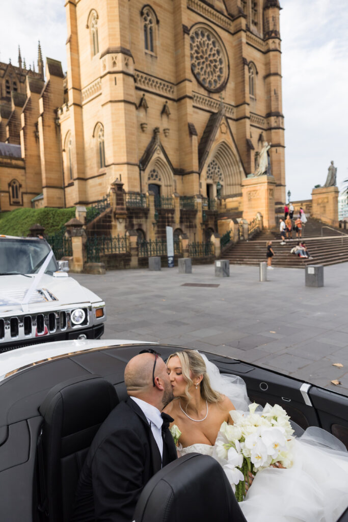 Bride and groom kiss in an open-top white car parked in front of a grand sandstone cathedral with steps and statues behind them, wedding scene