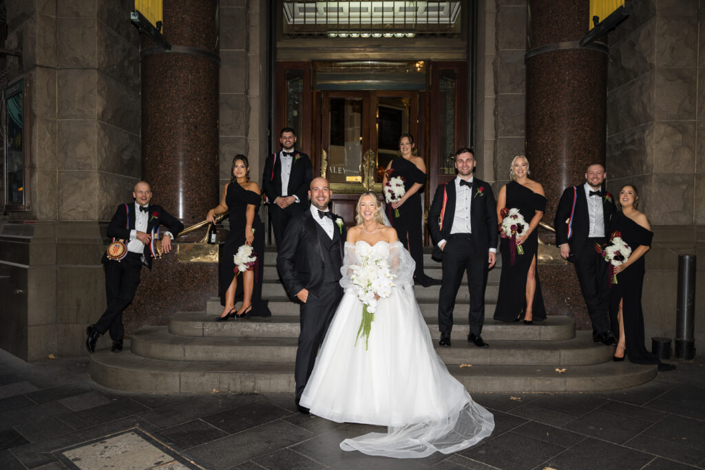 Bride and groom stand center on stone steps in formal wedding attire, surrounded by bridesmaids and groomsmen in black dresses and tuxedos outside a grand building.