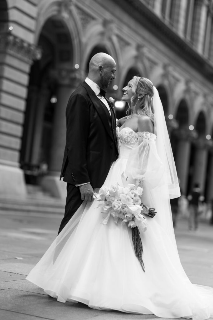 Bride in a white wedding gown and veil, holding a bouquet, smiling at her groom in a dark suit with arches of a building behind them.