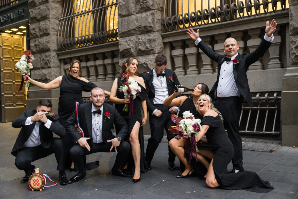 Group of formally dressed men and women posing playfully on a city street, holding bouquets and a bottle of champagne nearby.