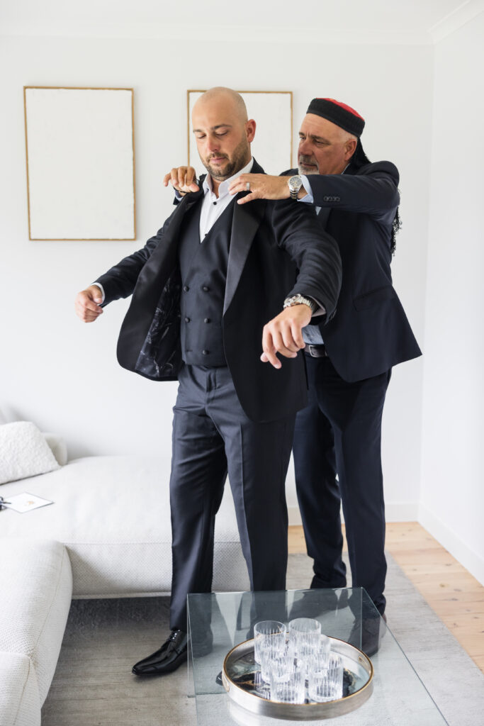 Two men in dark suits adjust each other’s jackets in a bright living room.
