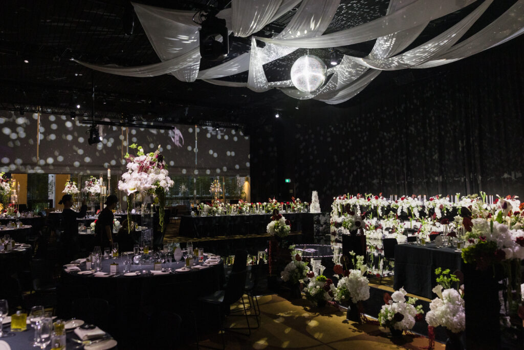 Banquet hall set for a formal event with long dining tables, tall floral centerpieces, and a sparkling disco ball under white draped ceiling fabric.