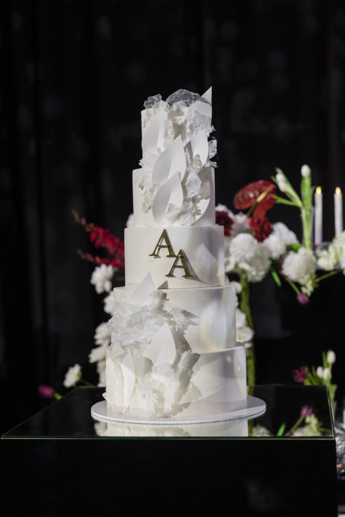 Tall white multi-tier cake adorned with crystal shards and white leaf shapes, featuring gold 'AA' initials on the middle tier, with floral decor in the background.