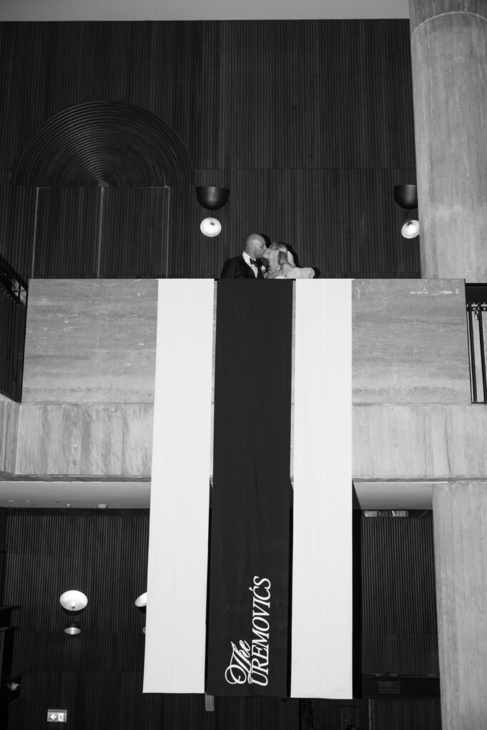 Bride and groom kiss on a balcony beneath vertical banners inside a modern building, celebrating a wedding moment.