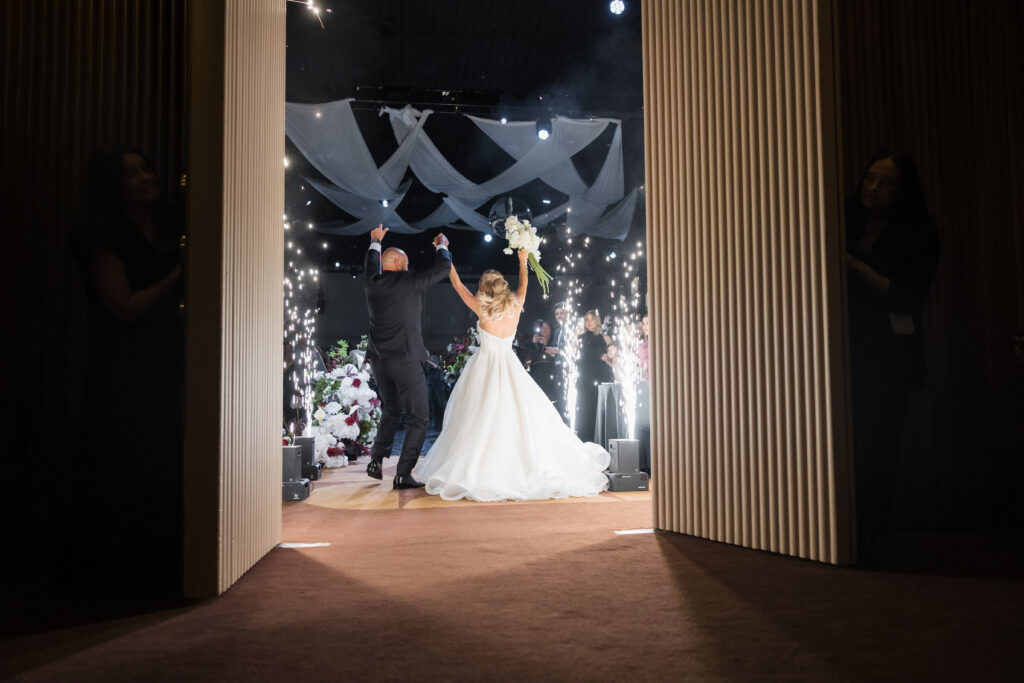 Bride in a white wedding gown and groom in a dark suit raise their arms in celebration as guests cheer behind them, framed by doorway and hanging fabric.