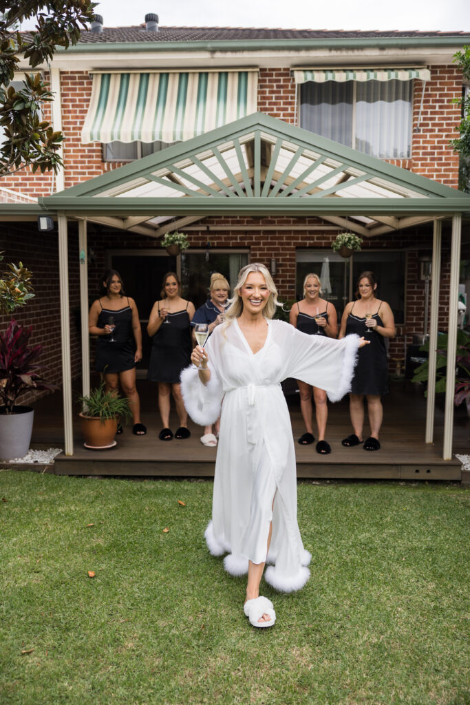 Bride in a white robe with fluffy cuffs walks toward the camera on the lawn, while bridesmaids in black dresses hold champagne glasses behind her.