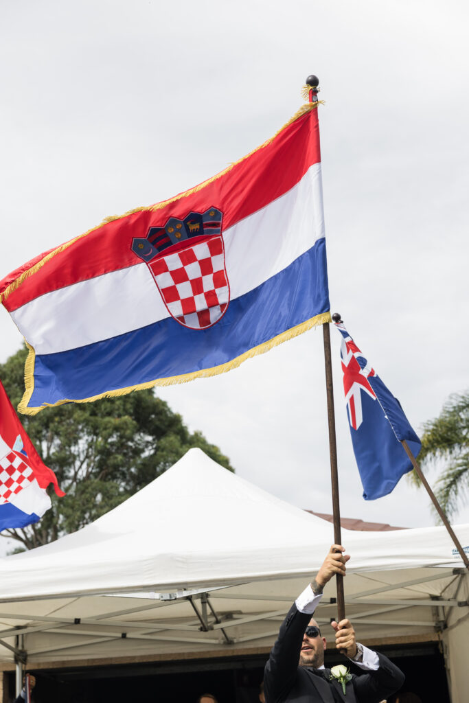 Croatian flag with red-white-blue stripes and crest waves outdoors, held by a person in a suit under tents.