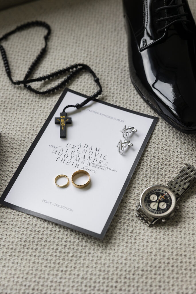 Close-up of wedding items on a carpet: black patent shoe, rosary-like necklace with cross, rings, cufflinks, and a printed invitation card with a watch nearby.