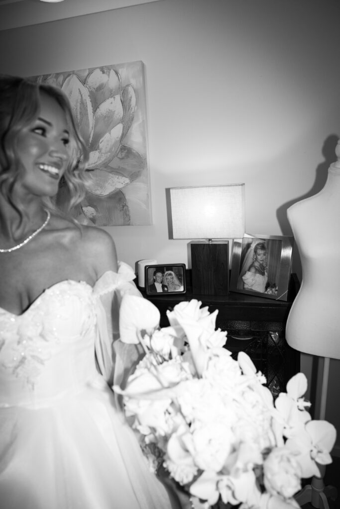 Bride in a wedding dress smiling, holding a bouquet of white flowers in the foreground; bedside table with framed photos and a lamp in the background.”]} }{