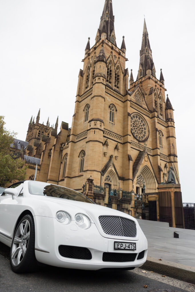 White Bentley parked on a street with a Gothic cathedral visible in the background.