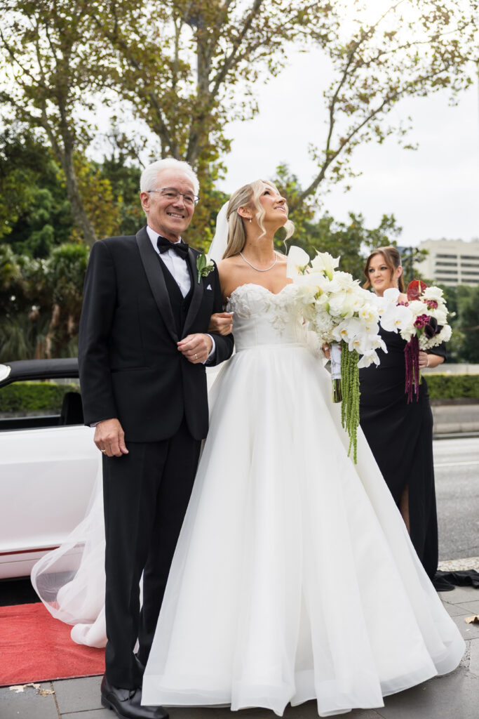 Bride in a white wedding gown stands beside an older man in a black tuxedo on a red carpet, with a bridesmaid holding a bouquet in the background.