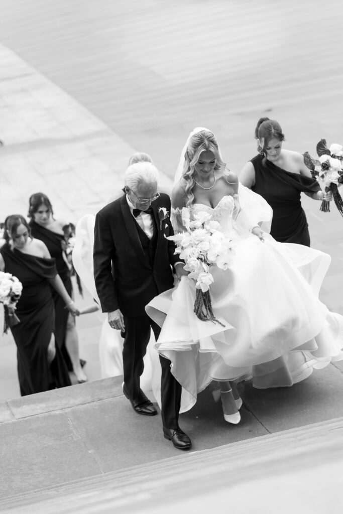 Bride in a white wedding gown descending stone stairs, escorted by a man in a suit, with bridesmaids in black dresses following and holding bouquets.