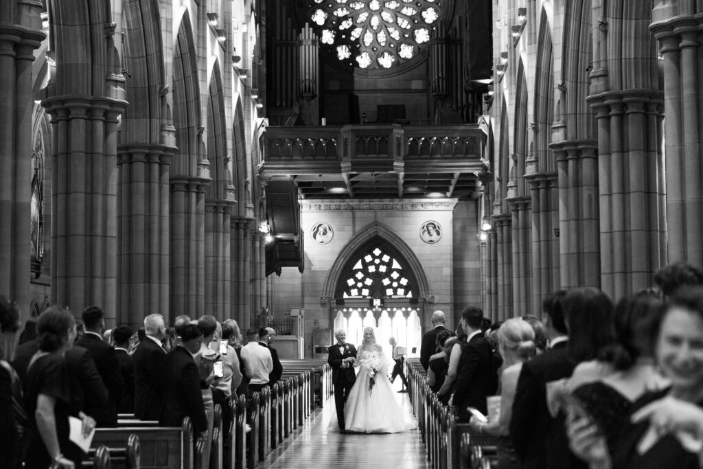 Bride and groom walk down the church aisle during a wedding ceremony, flanked by seated guests in a Gothic cathedral.