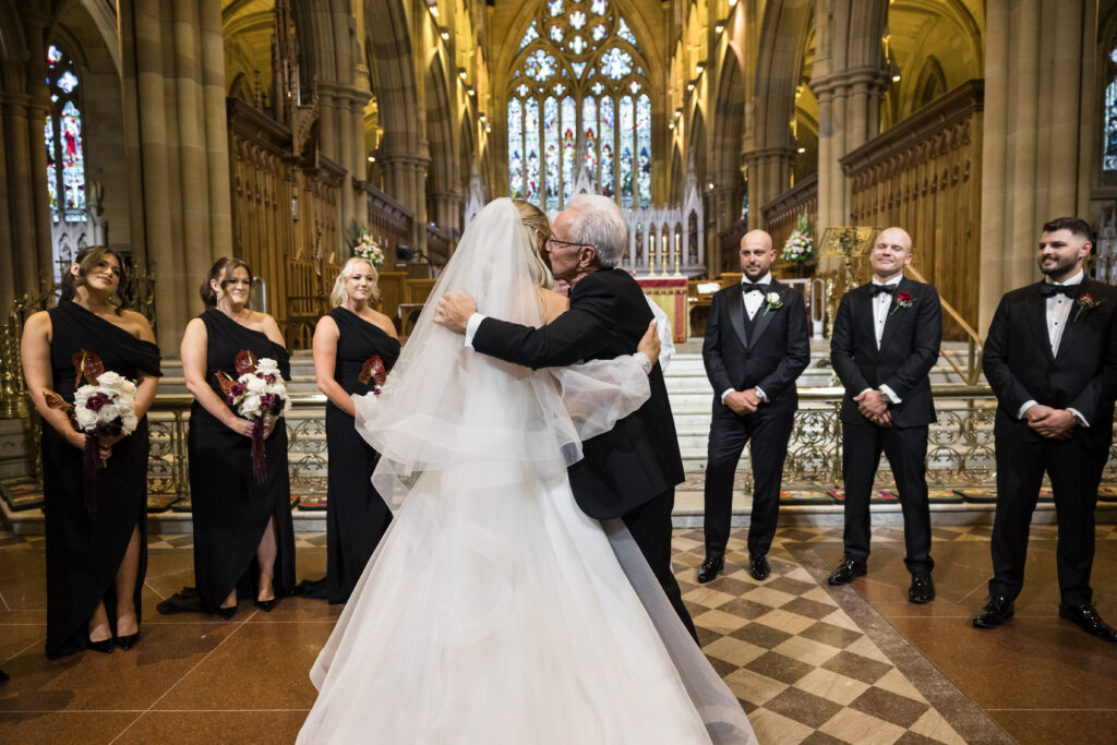 Bride in a white gown hugs an elderly man at the altar while bridesmaids in black dresses hold bouquets and groomsmen in tuxedos watch in a grand church with stained glass behind them, during a wedding ceremony.
