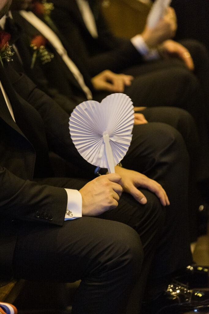 Person in a dark suit seated, holding a white pleated handheld fan in front of their lap at an event
