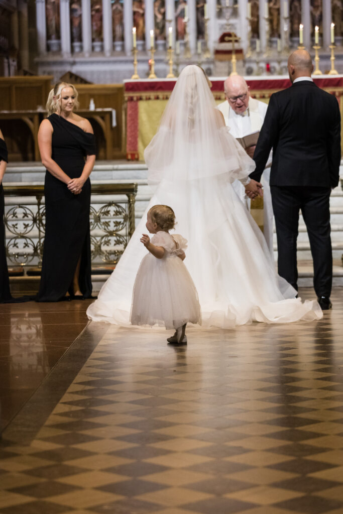 Bride in a white gown and veil at the altar with the groom; a small child in a white dress walks toward them in the foreground inside a church.