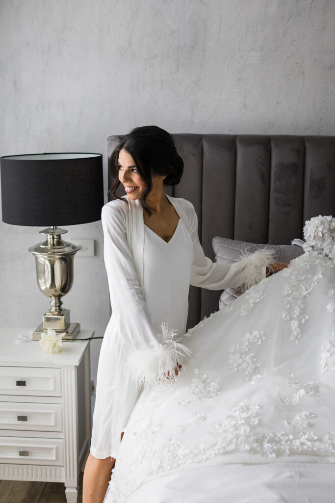 Woman in a white robe stands beside a bed, adjusting a lace wedding gown with floral appliqués and feathered cuffs.