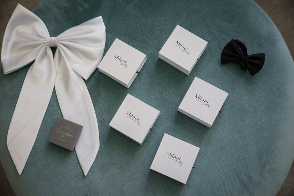 Milani Jewelry white boxes arranged on a teal surface with a large white satin bow on the left and a black bow on the right.