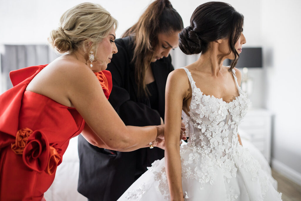Bride in an intricate white lace wedding dress stands in profile as two helpers assist with the back, in a bright room.