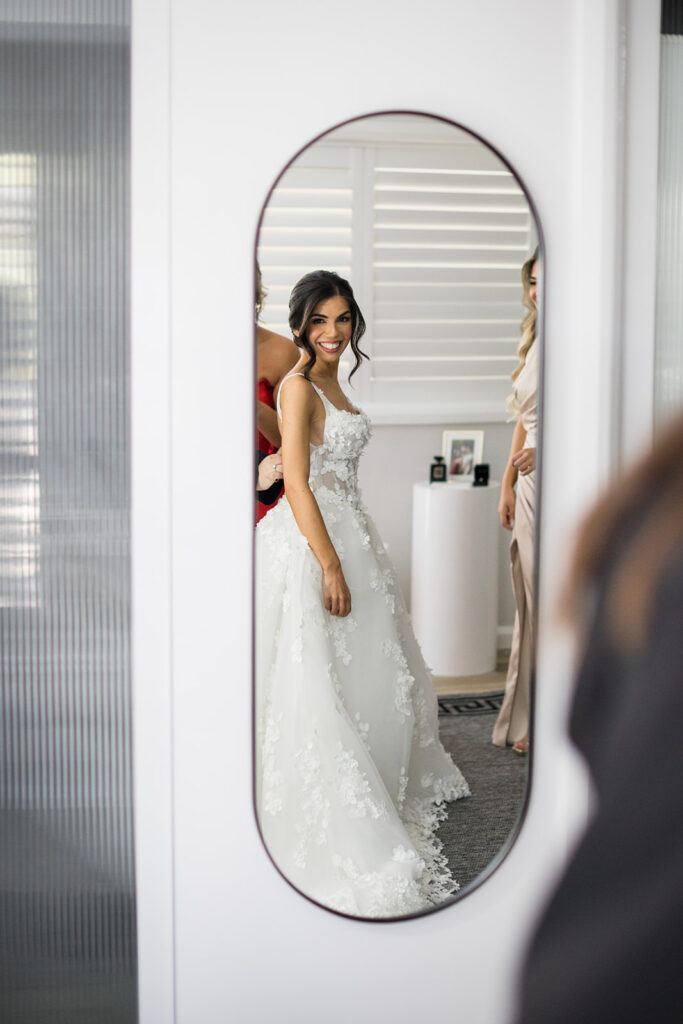 Bride in a lace wedding dress smiles at her reflection in an oval mirror.