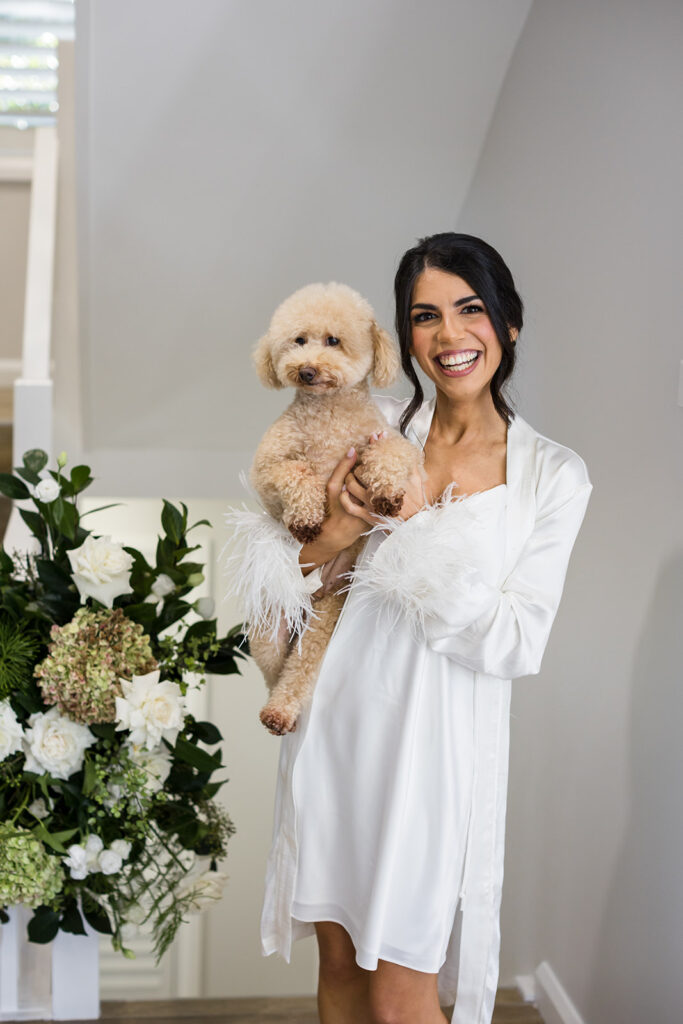 Smiling woman in a white robe holding a fluffy apricot poodle indoors, with a floral arrangement nearby.