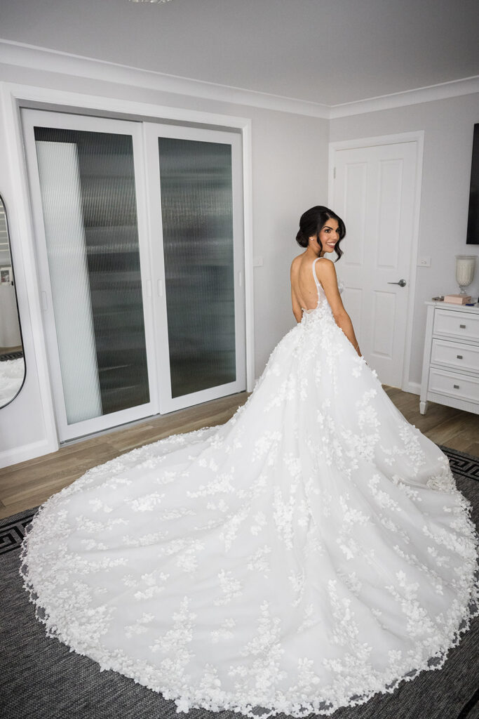 Bride in a white floral lace wedding gown with a long train, standing in a bright room and smiling back over her shoulder.