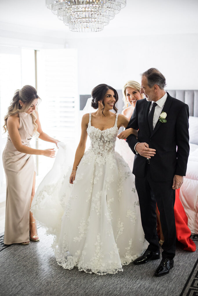 Bride in a lace wedding gown smiles as a man in a black suit helps her with her dress, while two bridesmaids stand nearby in a bright room.