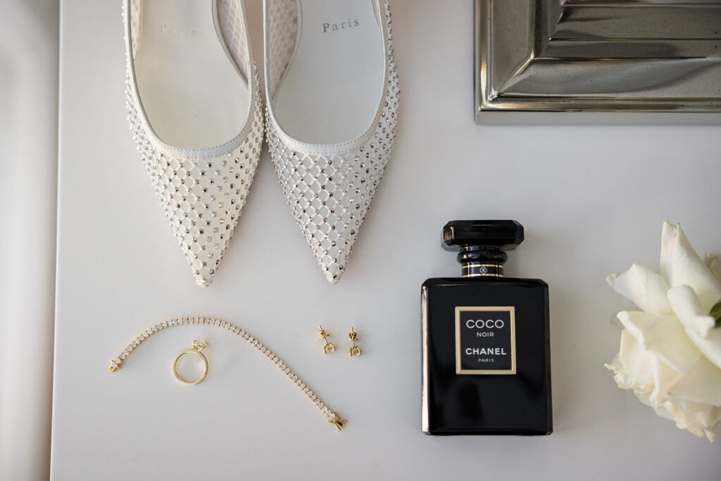 White rhinestone‑embellished pointed flats paired with jewelry and a Chanel Coco Noir perfume bottle on a white surface, with a white rose in the corner.
