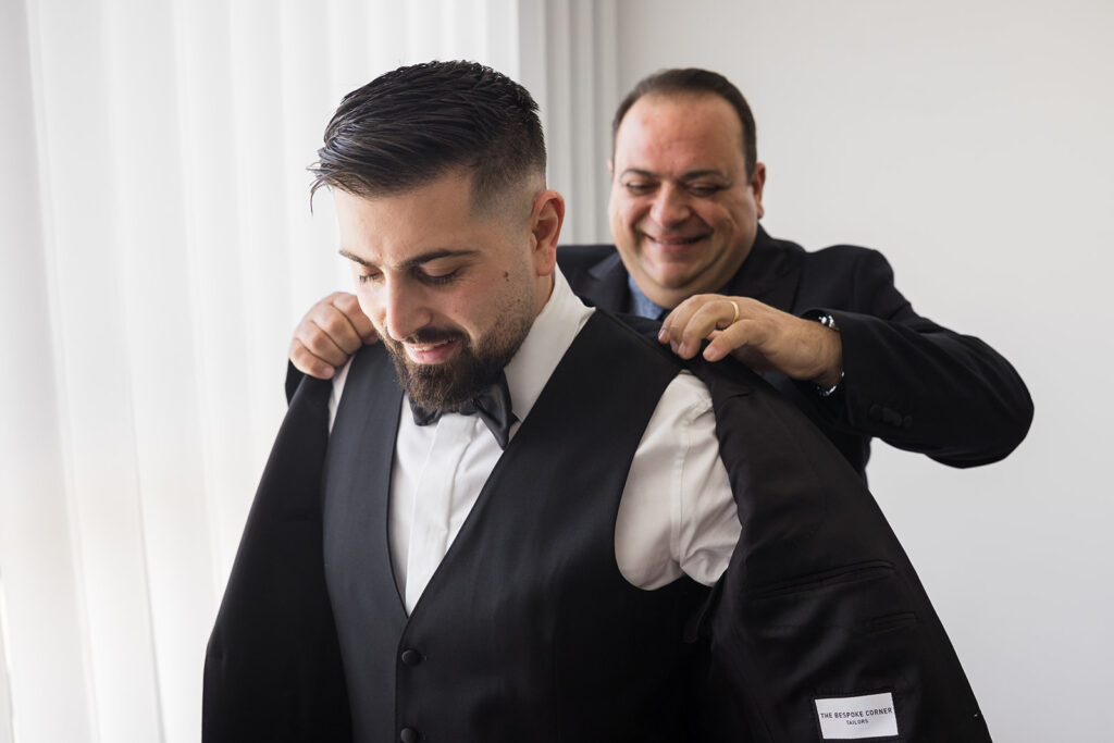 Groom getting helped into a black suit jacket by a smiling tailor in a dark suit, indoors beside white curtains.