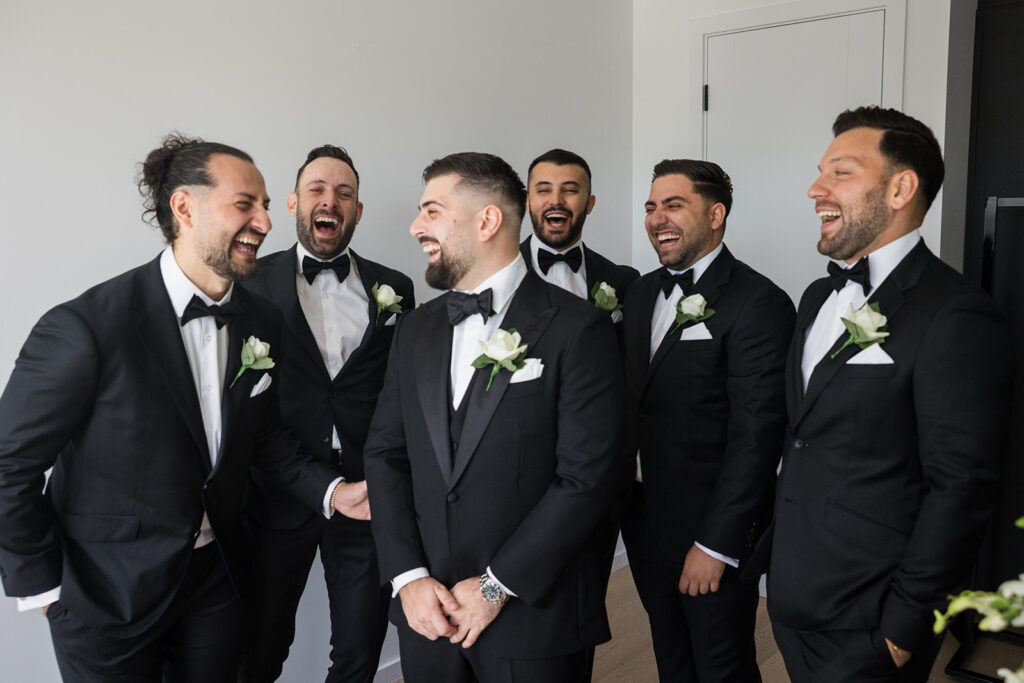 Six men in black tuxedos with bow ties laugh together in a bright room, sharing a joyful moment.
