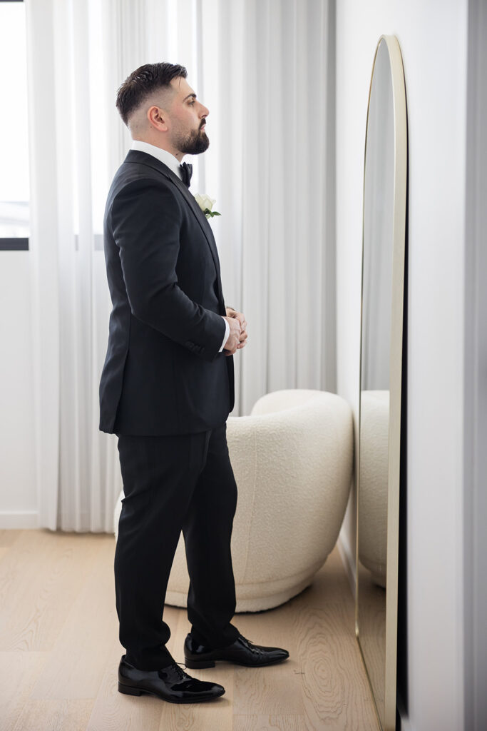 Man in a black tuxedo with a white boutonniere stands facing a tall mirror in a bright room, preparing for a formal event.