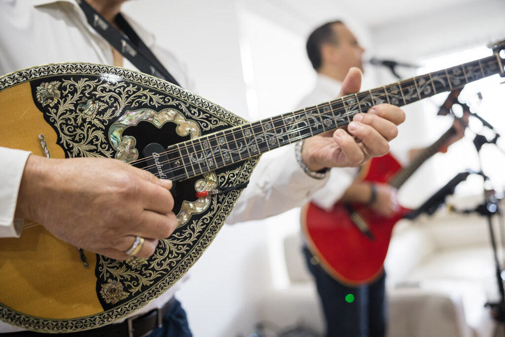 Close-up of a musician's hands playing an ornate mandolin with intricate floral inlay and pearl accents on a black pickguard; focus on strings and frets.
