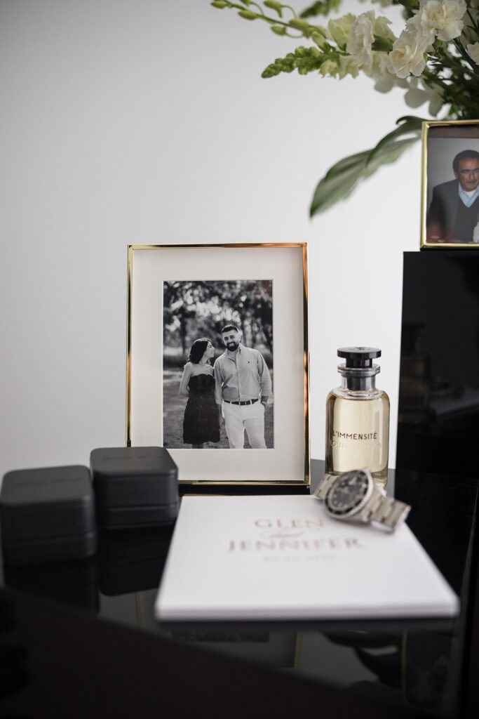 Gold-framed black-and-white photo of a couple on a glossy desk, with a bottle labeled L'IMMENSITÉ nearby and a silver watch nearby.
