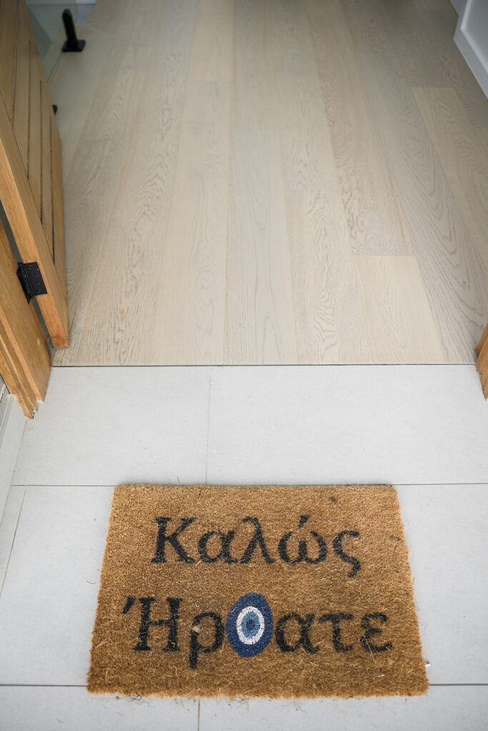 Brown coir welcome mat at doorway with Greek text 'Καλώς Ηρθατε' on light tile floor outside room.