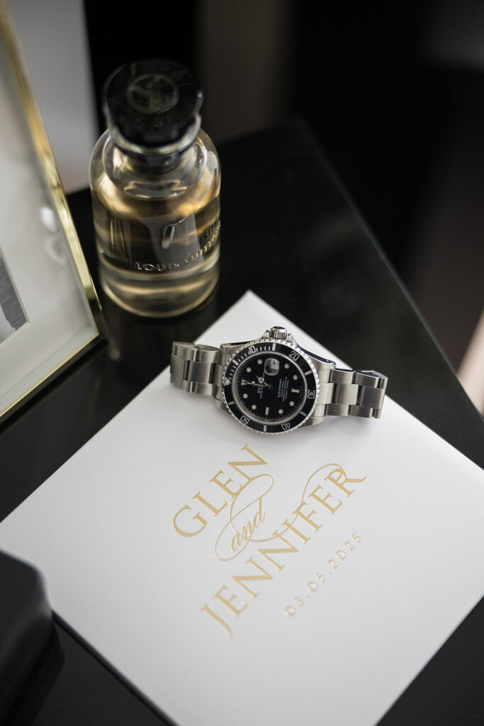 Silver stainless steel wristwatch resting on a white wedding invitation card with gold text reading 'GLEN and JENNER 03.05.2023', a perfume bottle visible in the background.