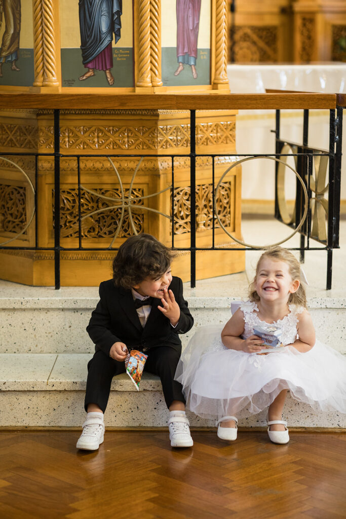 Two young children in formal clothes sit on church steps, smiling—girl in a white dress, boy in a black suit eating a snack.