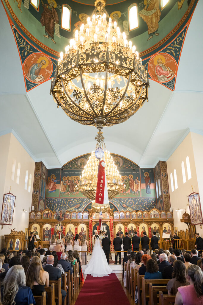 Bride and groom stand at the ornate iconostasis during a wedding ceremony inside an Orthodox church, with guests seated in pews and chandeliers overhead.