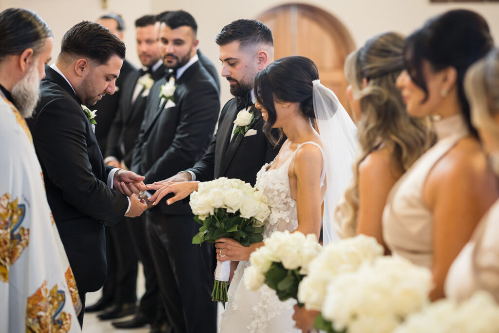 Bride and groom exchange rings during a wedding ceremony, surrounded by bridesmaids and groomsmen with white bouquets.