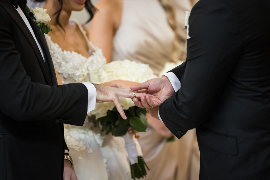 Two men in black suits exchange wedding rings, with a bride in a white lace dress and bouquet visible in the background.