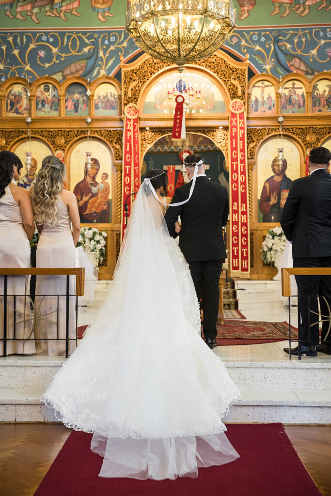 Bride and groom walk down the church aisle during a wedding ceremony, with bridesmaids in pale dresses nearby and an ornate icon wall behind them.