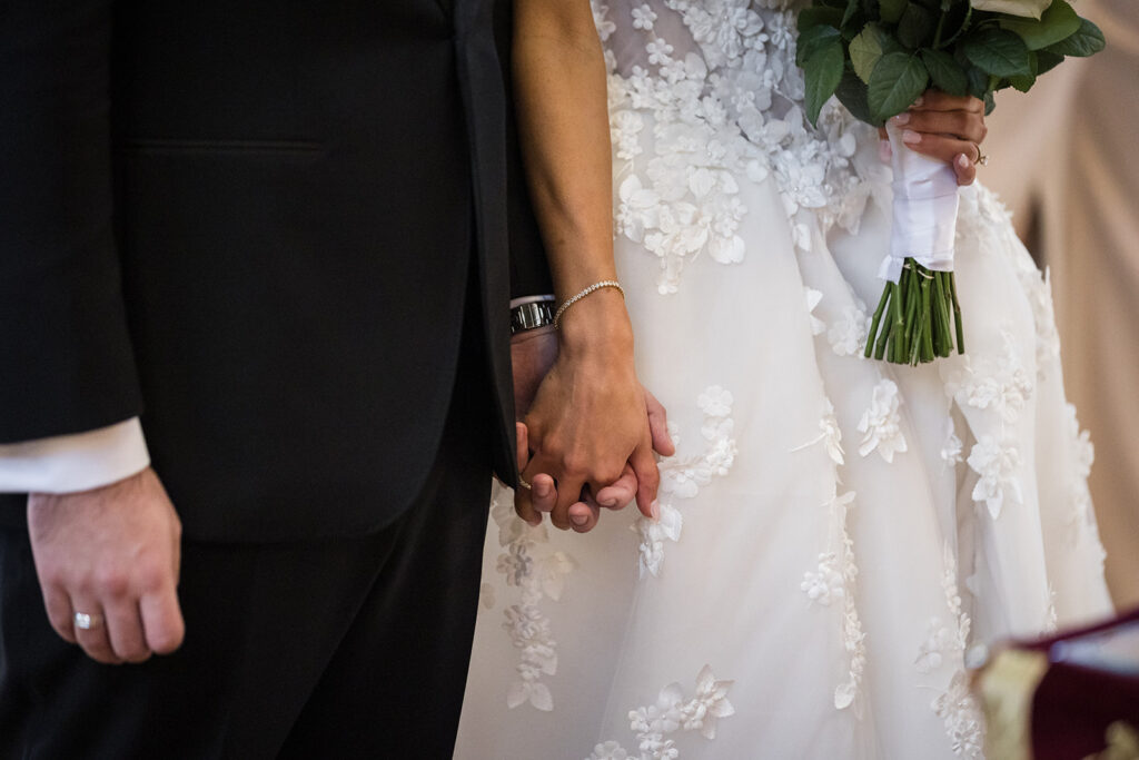 Bride and groom hold hands during a wedding; the bride wears a white lace gown and holds a bouquet with green stems.