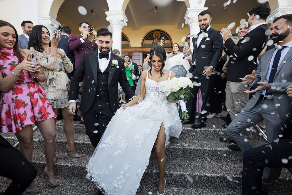 Bride in a white lace gown with a bouquet descends stone steps as groom in a black suit follows, guests tossing white confetti and photographing the moment outdoors at a wedding venue.