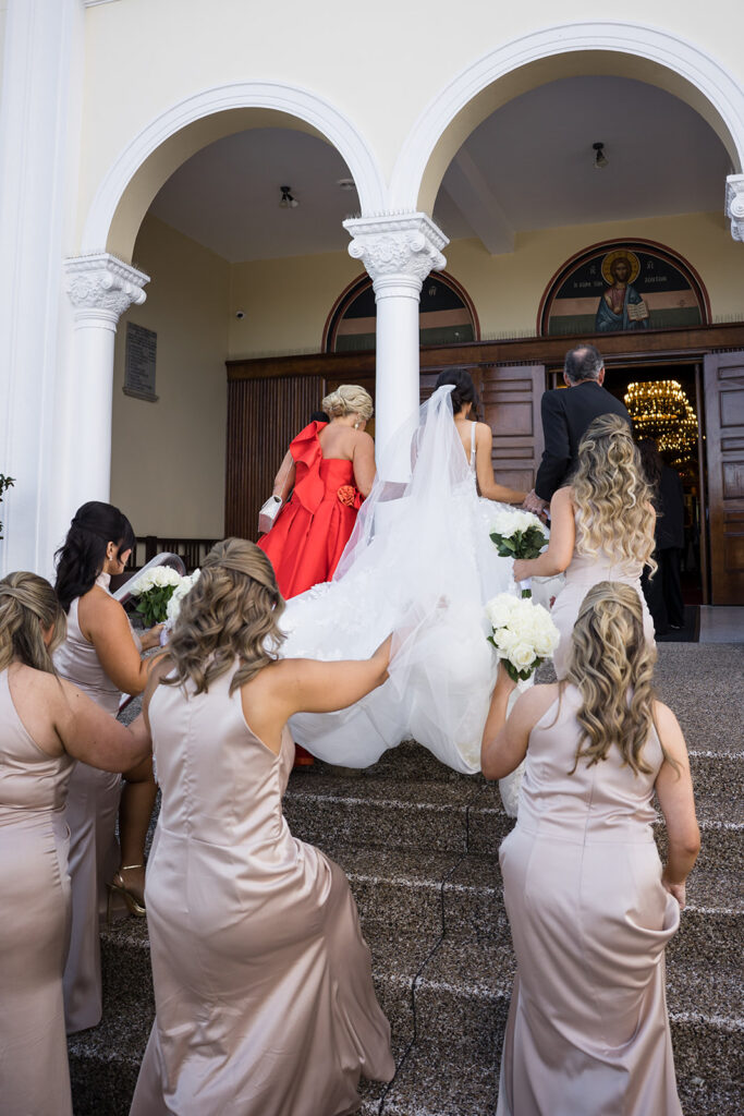 Bridesmaids in beige dresses assist the bride in a white gown as they walk up church stairs toward the open doors, holding white bouquets.