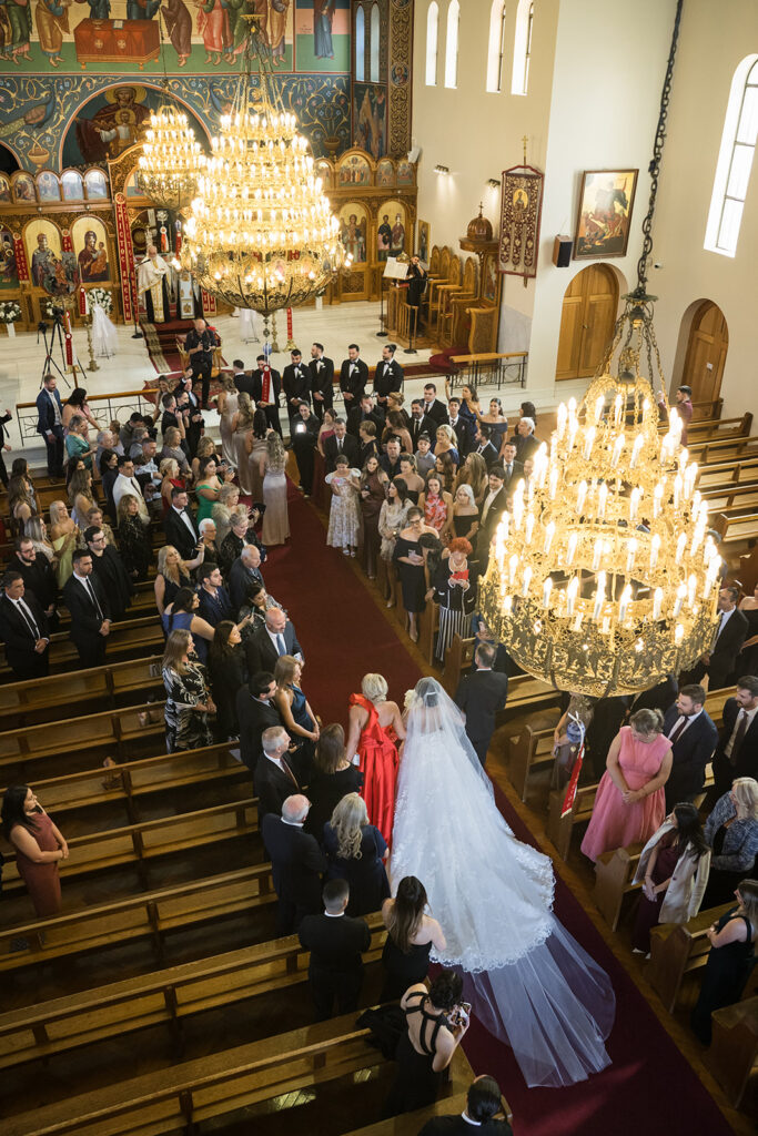 Bride in a white wedding gown with long veil walking down a red carpet aisle inside an ornate church, surrounded by formally dressed guests.